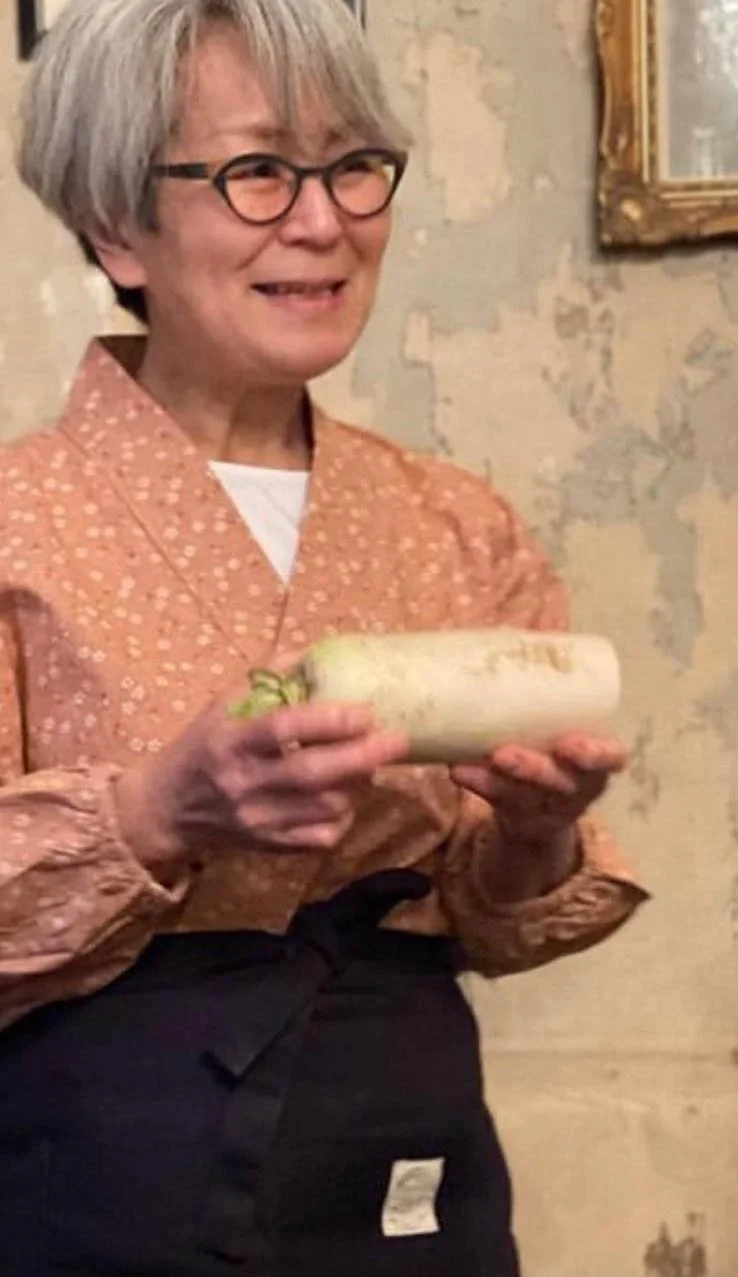 An older woman with gray hair and glasses holding a large daikon radish, smiling in front of a textured wall with a framed picture.