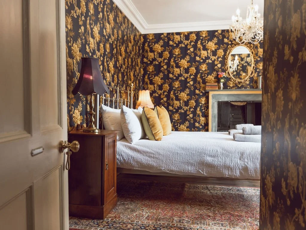 A cozy bedroom with dark floral wallpaper, a bed with white bedding and multiple pillows, two lamps, a mirror above the fireplace, a small table with a flower vase, and a hanging chandelier.