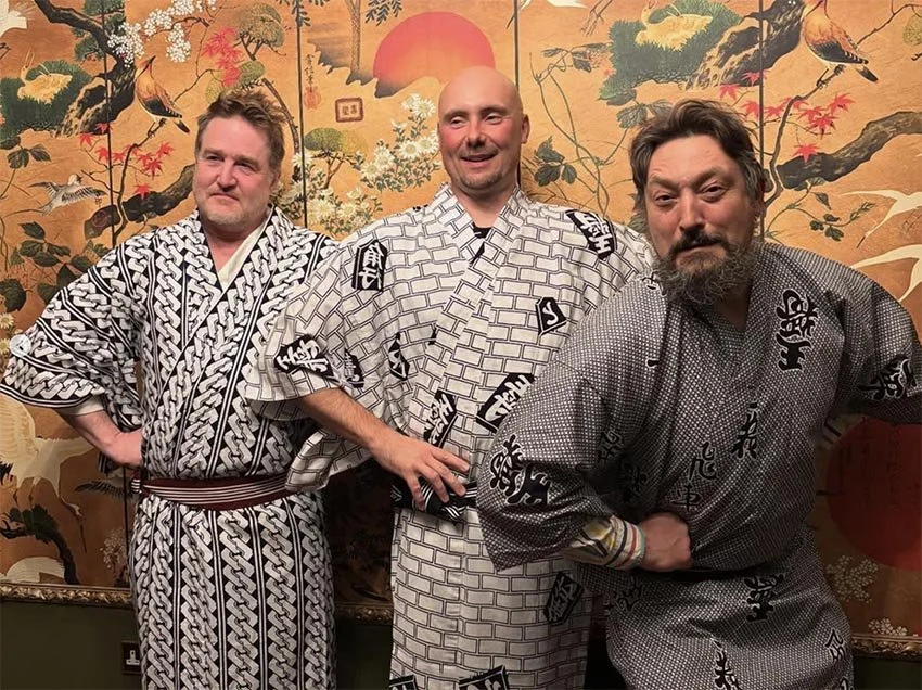 Three men dressed in traditional Japanese yukatas posing in front of a decorative screen with a sunset, trees, and birds.