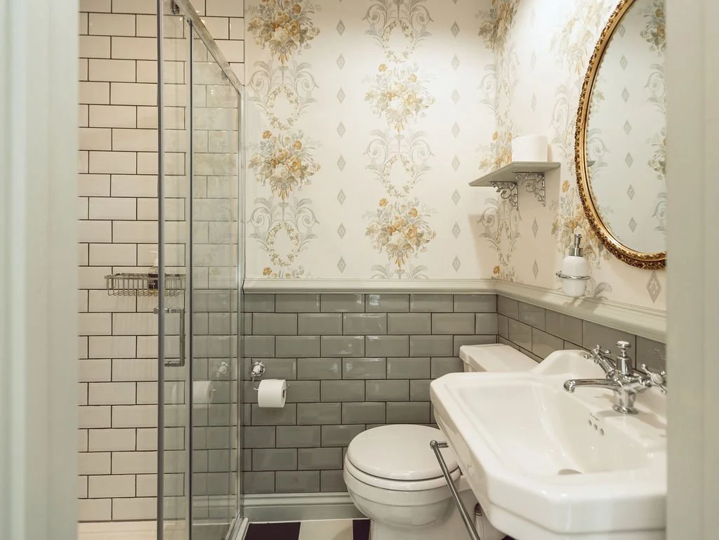 A small vintage-style bathroom with a shower stall, a toilet, and a white porcelain sink. The walls are decorated with floral wallpaper on the upper half and gray subway tiles on the lower half. There is a small shelf with a tissue box and a soap dis