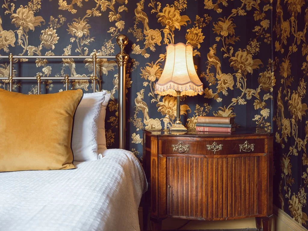 A bedroom corner featuring a bed with gold pillows, a vintage wooden nightstand with decorative brass handles, a table lamp with a fabric lampshade, and a floral wallpaper with gold and blue patterns.