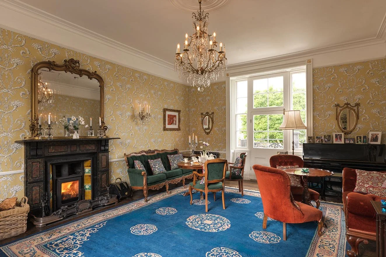 Elegant living room with patterned yellow wallpaper, a fireplace with a mirror above, a blue patterned rug, a green velvet sofa, four armchairs, a piano, and large windows letting in natural light.