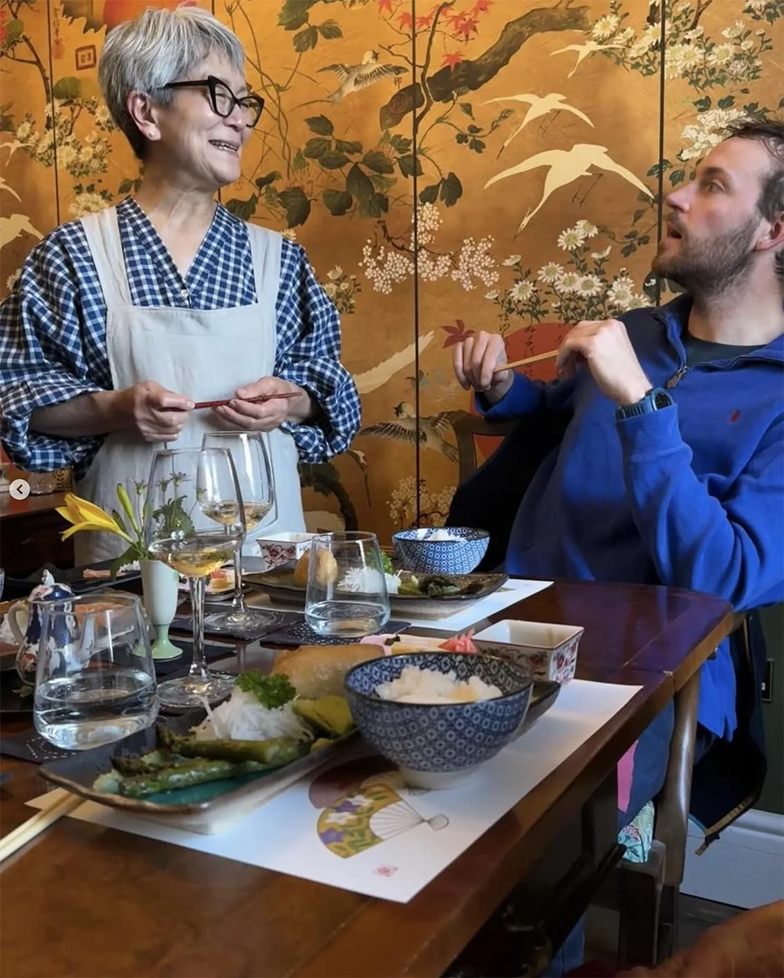 A woman with short gray hair, glasses, wearing a checked shirt and apron, stands at a dining table, smiling and holding chopsticks. A man in a blue jacket sits at the table, looking up at her, also holding chopsticks. The table is set with various Ja
