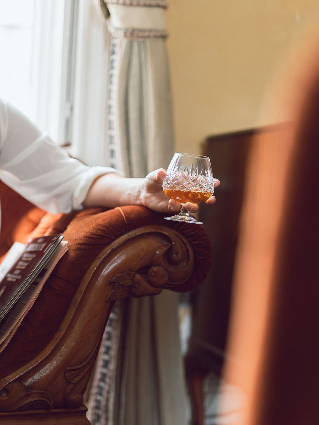 A person wearing a white shirt is holding a glass of whiskey in a carved wooden armrest of a sofa or chair in a room with beige wall and curtains.