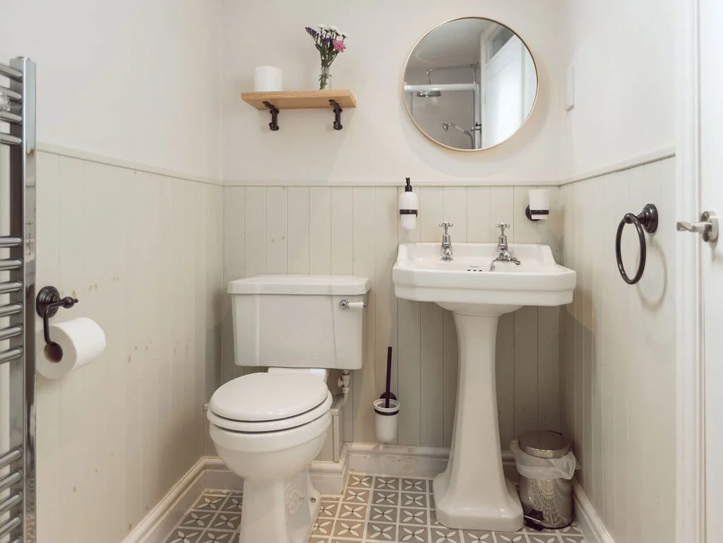 Small bathroom with white toilet, pedestal sink, round mirror, wall shelf with flowers, and patterned floor tiles.