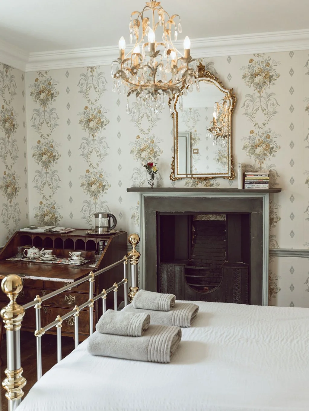 A cozy bedroom with a white bed, three rolled towels, a dresser with a tea set, a fireplace with a mirror above it, floral wallpaper, and a chandelier.