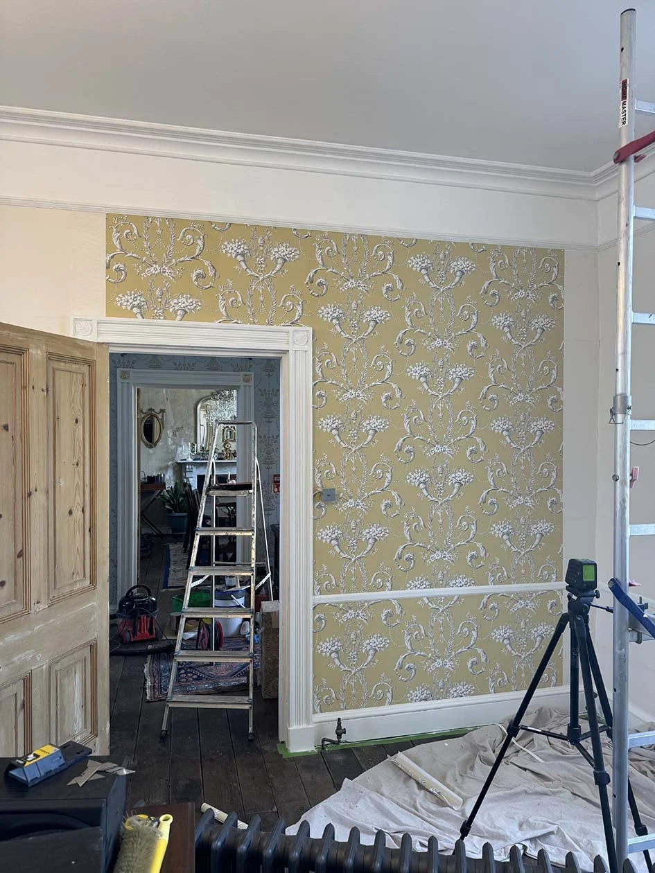 Interior of a home under renovation with a patterned yellow wallpapered wall, a tall stepladder, a tripod, and renovation tools and supplies in the room.