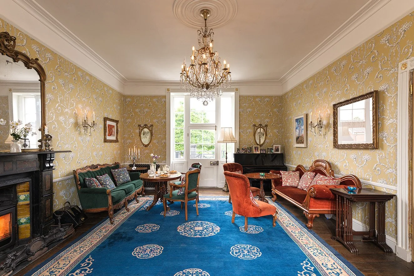 Living room with ornate chandelier, vintage furniture, fireplace with mirror, blue patterned rug, wall art, and large window with view outside.