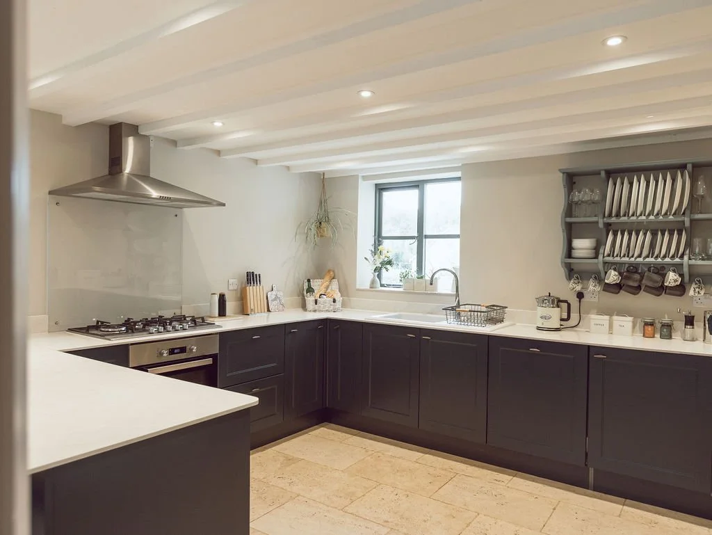 Modern kitchen with dark lower cabinets, white countertops, a window above the sink, open shelves with dishes, a gas stove with a range hood, and various kitchen appliances and utensils.
