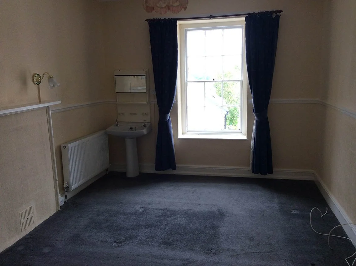 Empty room with a window, blue curtains, a wall-mounted mirror, a small sink, and a ceiling light fixture.