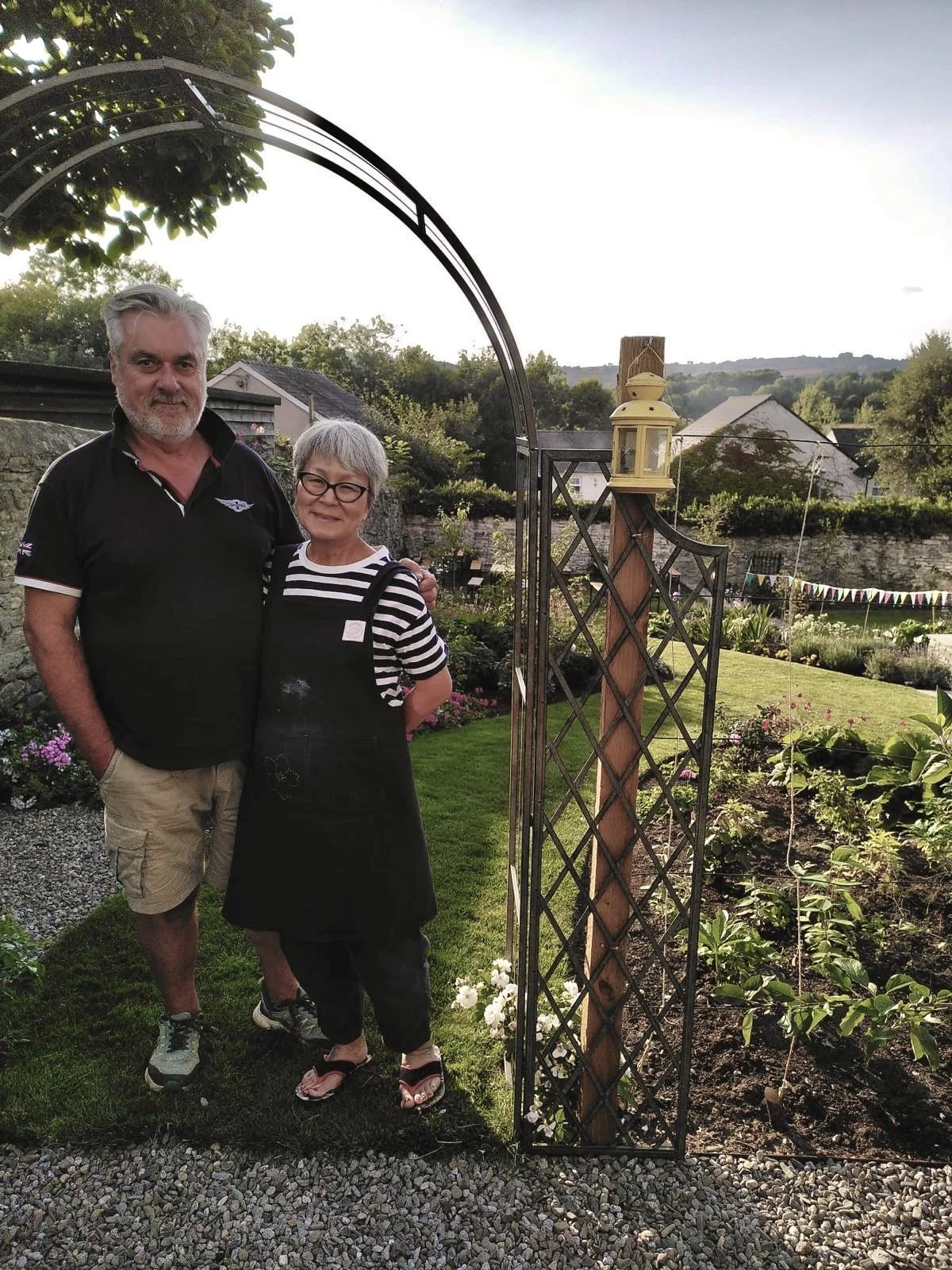 A man and woman standing together in a garden, posing near an arched metal gate with a yellow lantern on top, surrounded by plants and flowers, with houses and hills in the background during sunset.