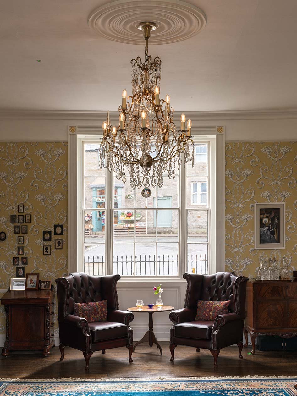 Interior of a sitting room featuring two dark leather armchairs, a small round wooden table with a small vase, glasses, and a bowl, a large window with a view of a street outside, a chandelier hanging from the ceiling, patterned wallpaper, framed pic