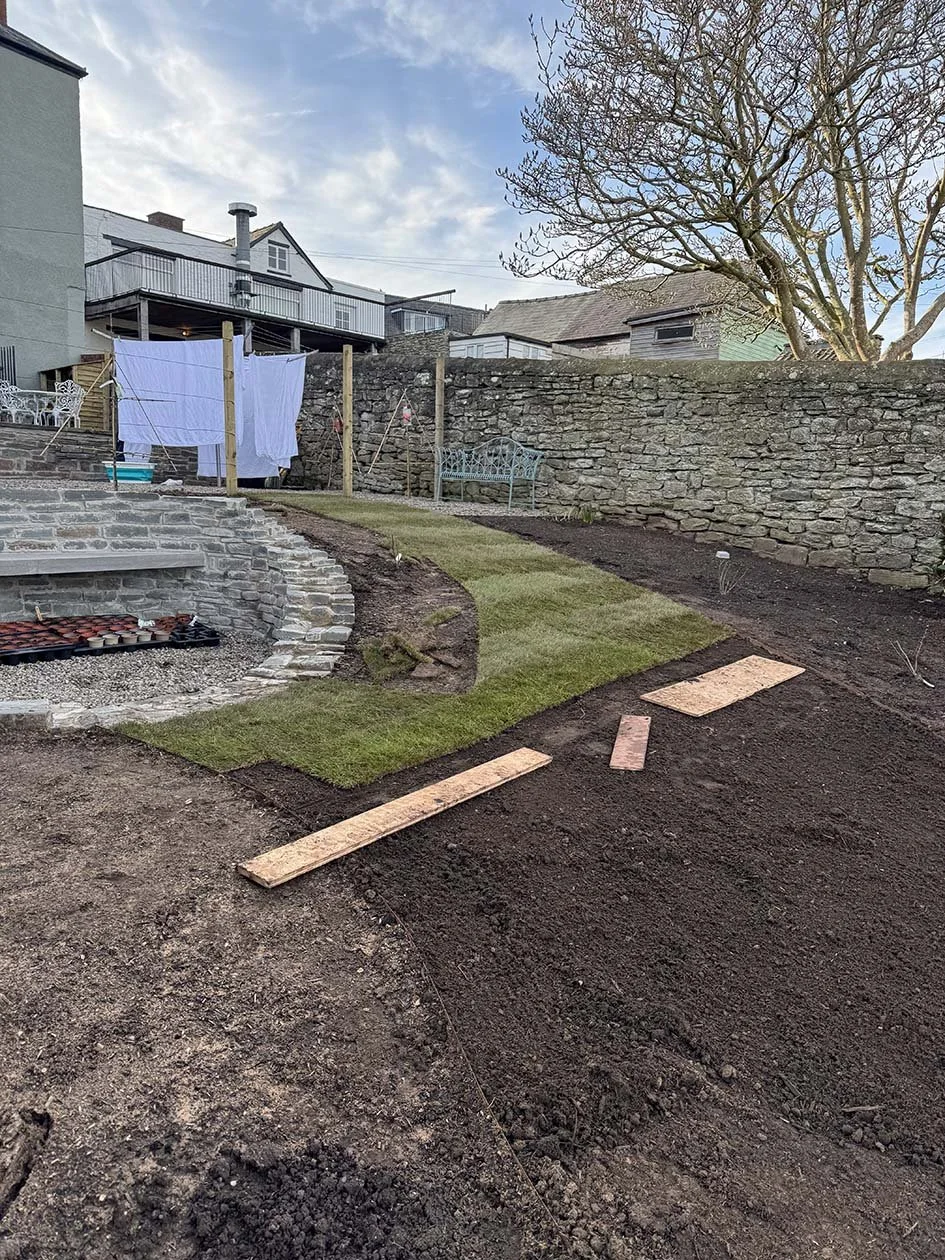 A backyard garden area with a newly laid patch of green grass, a stone fire pit, laundry hanging on clotheslines, and a stone wall in the background. The sky is partly cloudy with patches of blue, and some wooden planks and a rectangle of plywood are