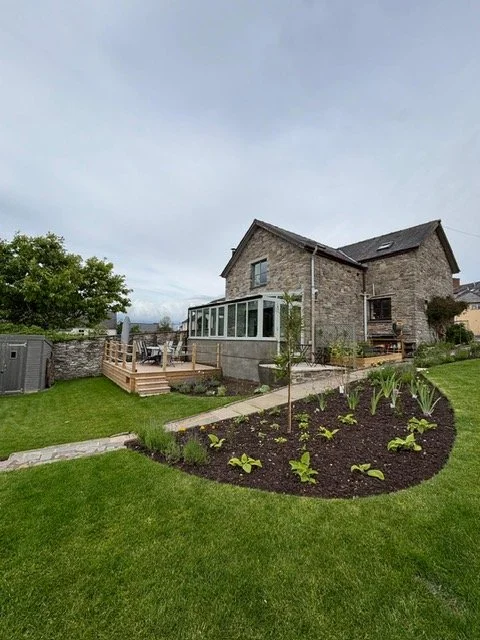 Backyard with a stone house, a wooden deck, and a garden with young plants and a small tree.
