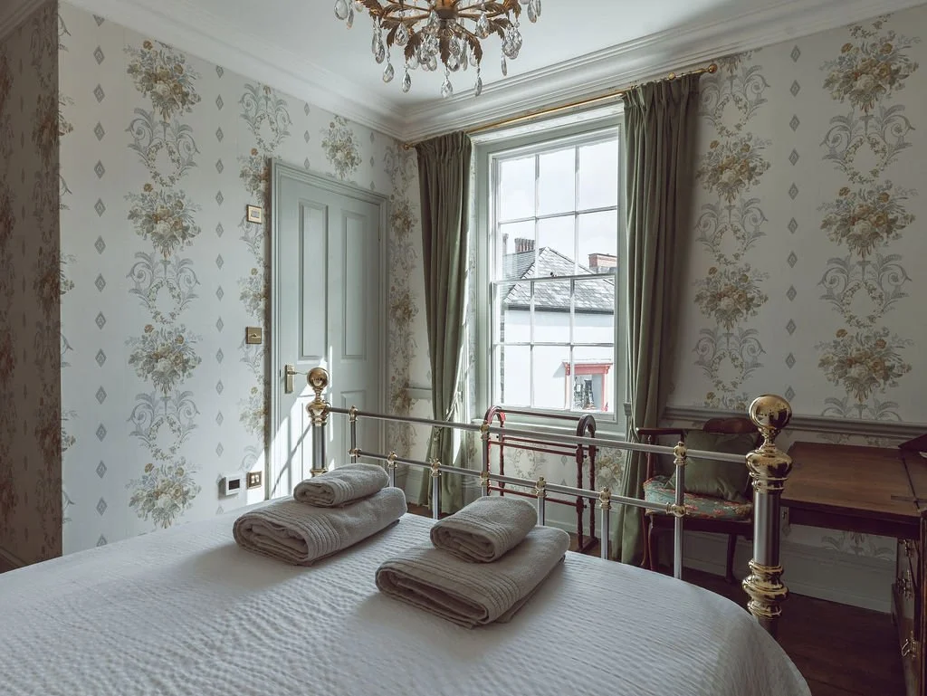 A cozy bedroom with a brass bed, floral wallpaper, a large window with green curtains, a chandelier, and a small wooden desk.
