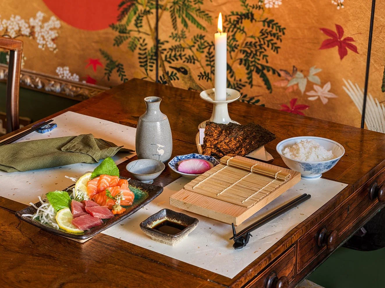 A traditional Japanese meal setup on a wooden table with sashimi, rice, seaweed, soy sauce, chopsticks, and a candle, with a decorative wall featuring Japanese motifs in the background.