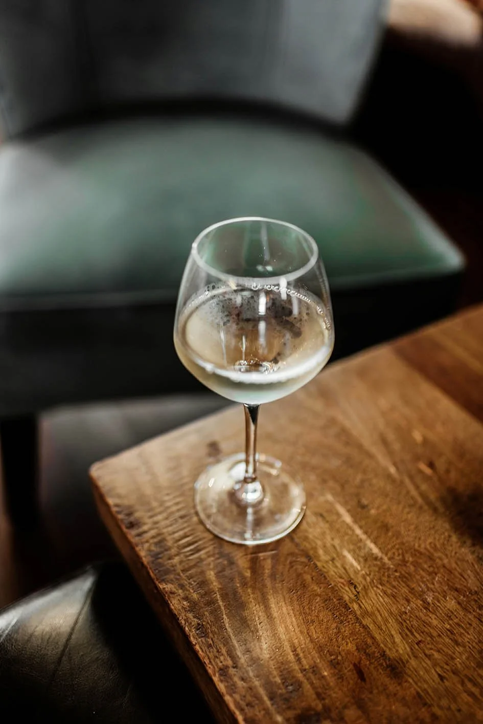 A glass of white wine or champagne on a wooden table, with a blurred green chair or seat in the background.