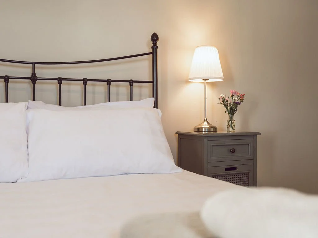 A neatly made bed with white pillows and bedding next to a nightstand with a lamp and a vase of pink flowers in a cozy bedroom.