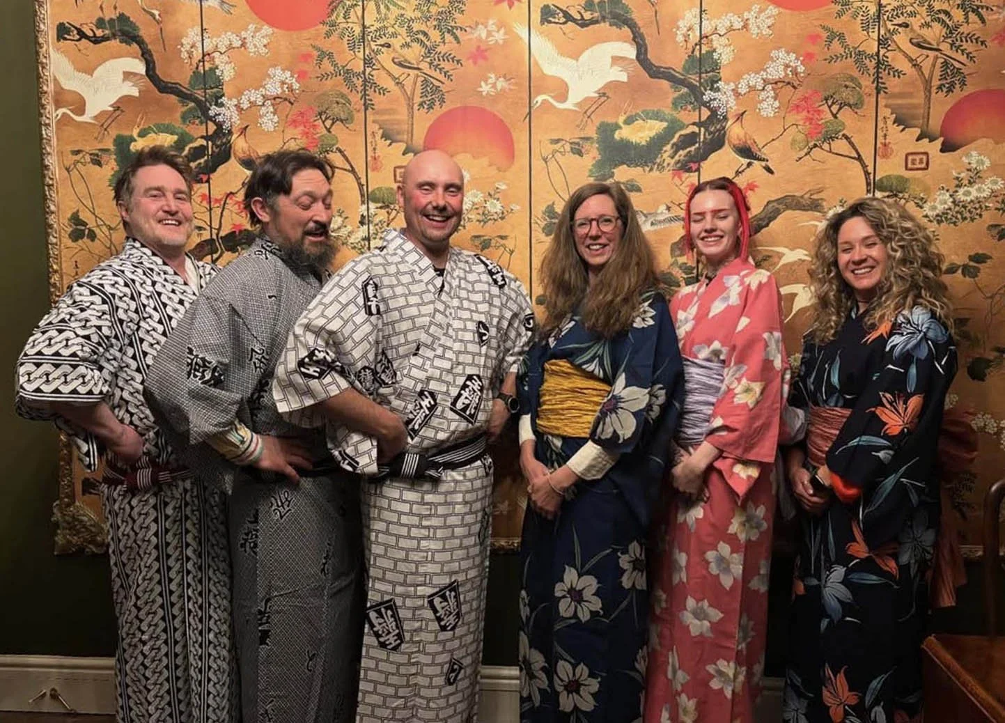 Group of six people wearing colorful and patterned yukatas, standing in front of a traditional Japanese screen with cranes, trees, and a setting sun.