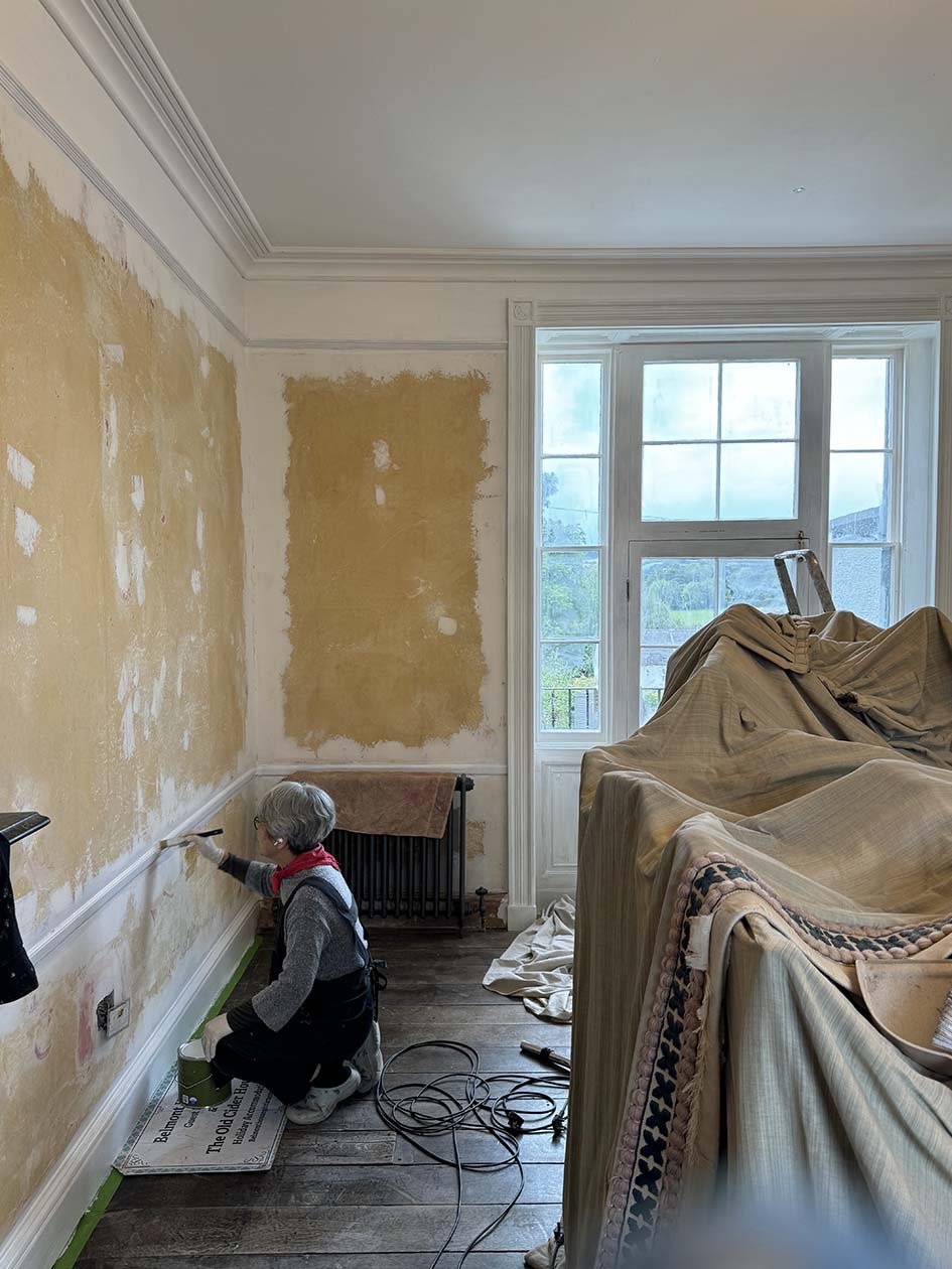 A person painting a wall in a room undergoing renovation, with furniture covered and a large window showing an outdoor view.