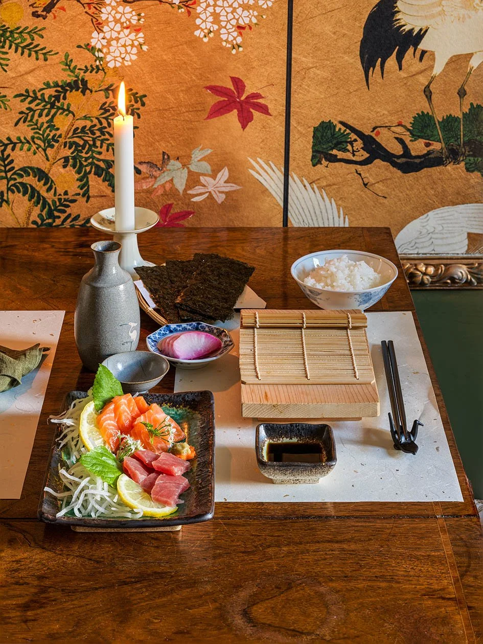 Japanese-themed dining setting with sashimi platter, rice bowl, seaweed sheets, soy sauce, chopsticks, and traditional decor background.