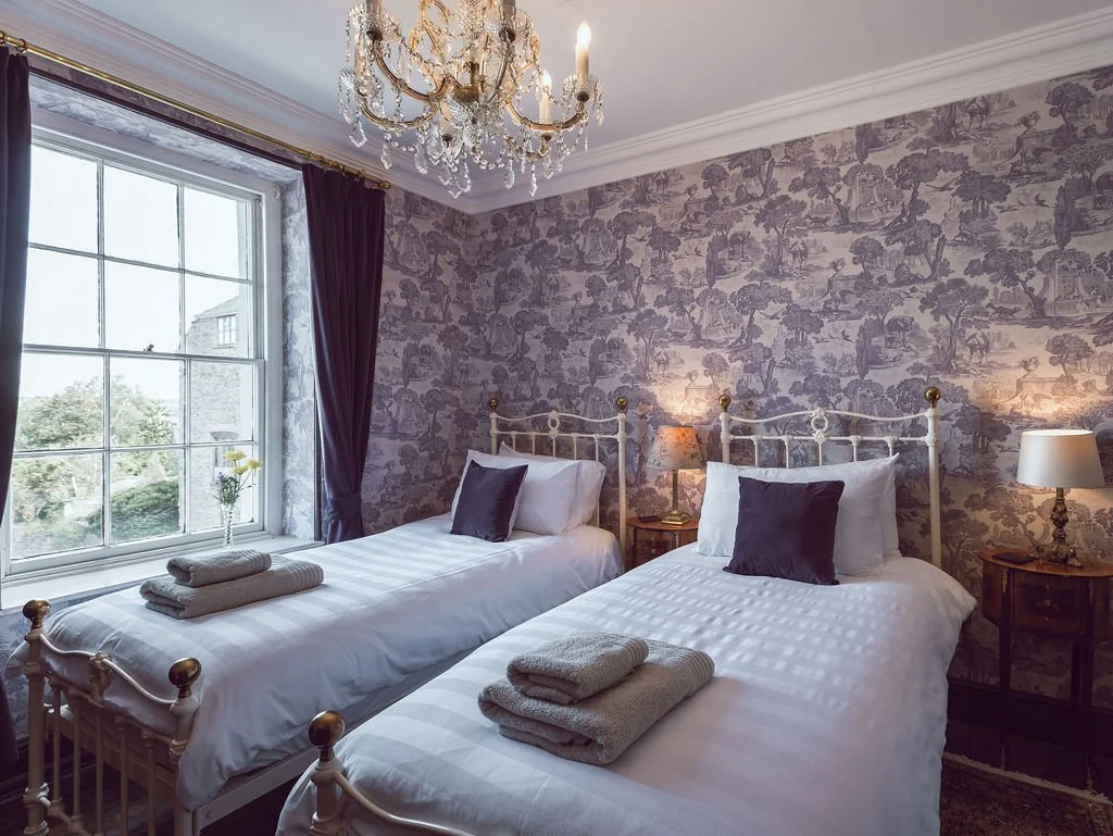 A bedroom with two single beds, white bedding, and dark pillows, placed against a patterned wallpapered wall. Each bed has neatly folded towels at the foot. There are bedside tables with lamps, and a window with purple curtains letting in natural lig