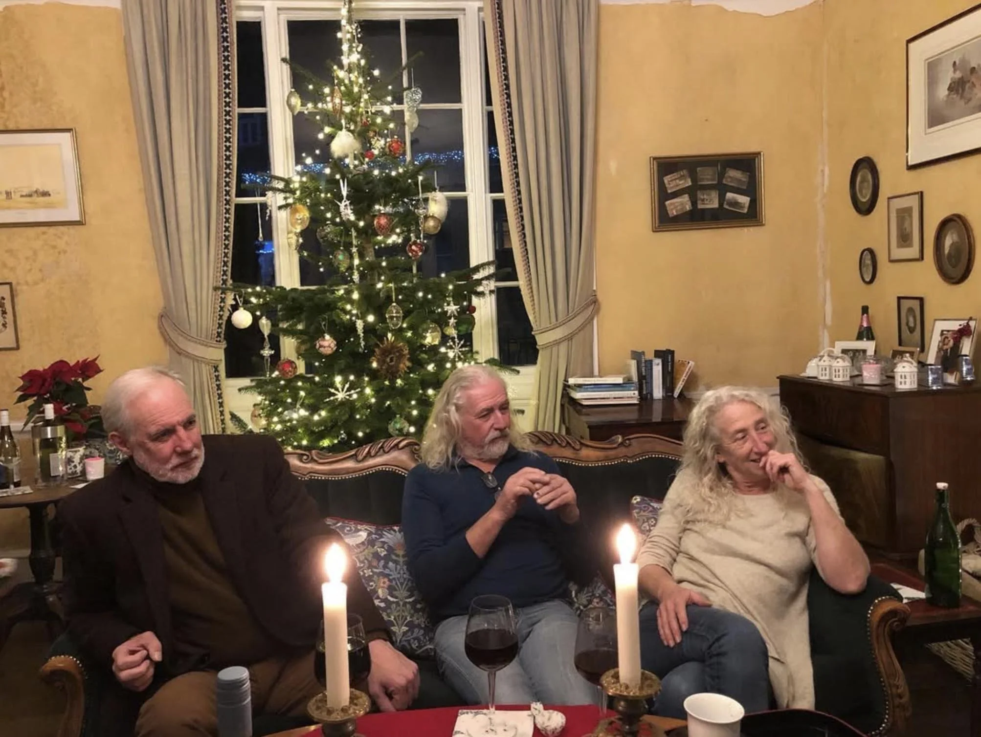 Three older adults, two men and one woman, sitting on a sofa in a living room decorated for Christmas with a lit Christmas tree and candles on the table.