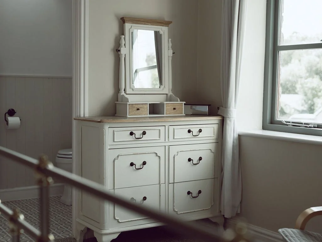 White vintage dresser with mirror in a bright room next to a window with white curtains.