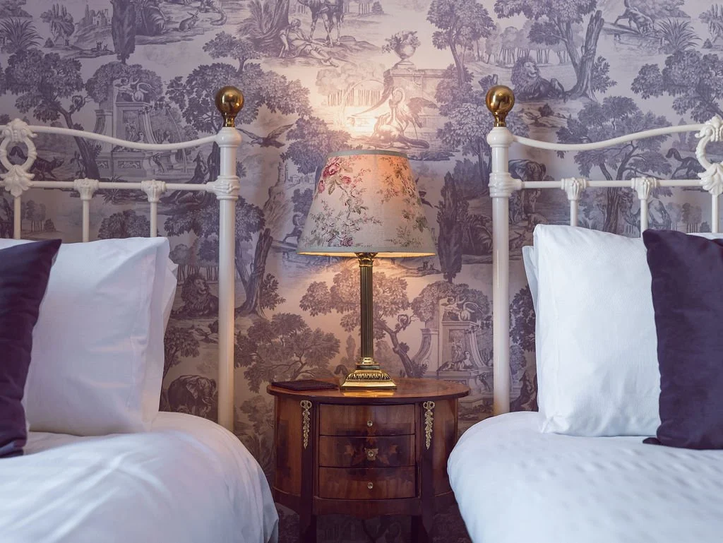 Two white beds with purple pillows flanking a wooden nightstand with a floral table lamp, against a blue and white scenic wallpaper in a vintage-style bedroom.