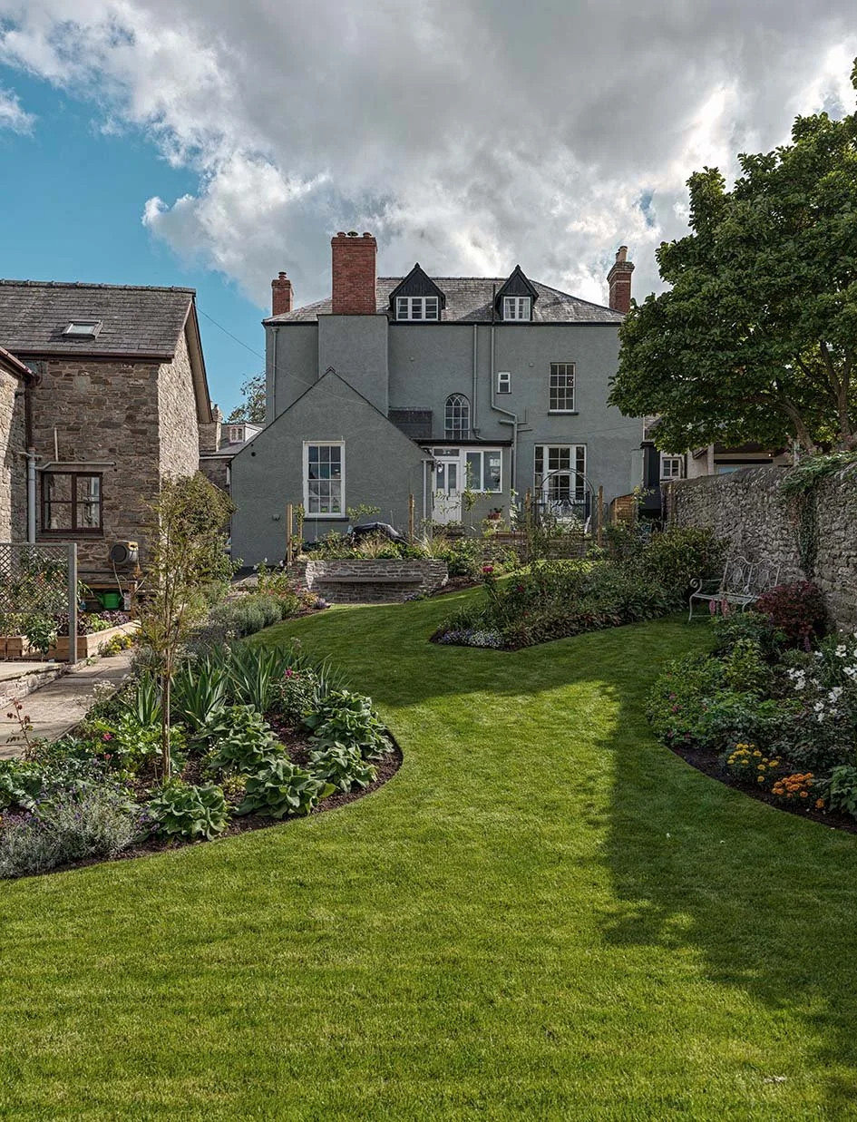 View of a backyard garden with a well-maintained lawn, garden beds with flowers and plants, a stone wall, a bench, and a tall gray house with red chimneys and dormer windows in the background under a partly cloudy sky.