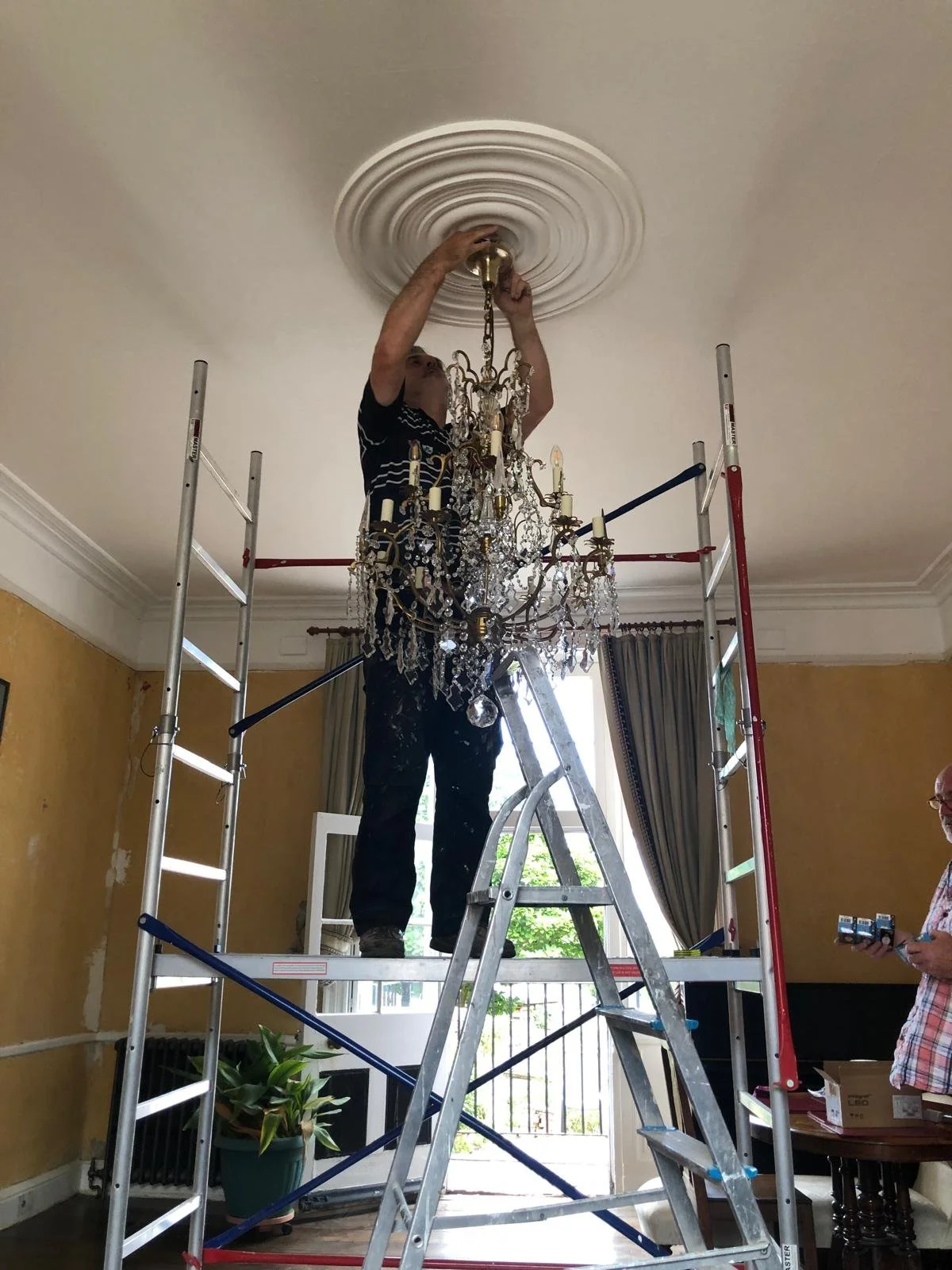A man on a ladder installing or fixing a chandelier on the ceiling of a living room, with another person observing.