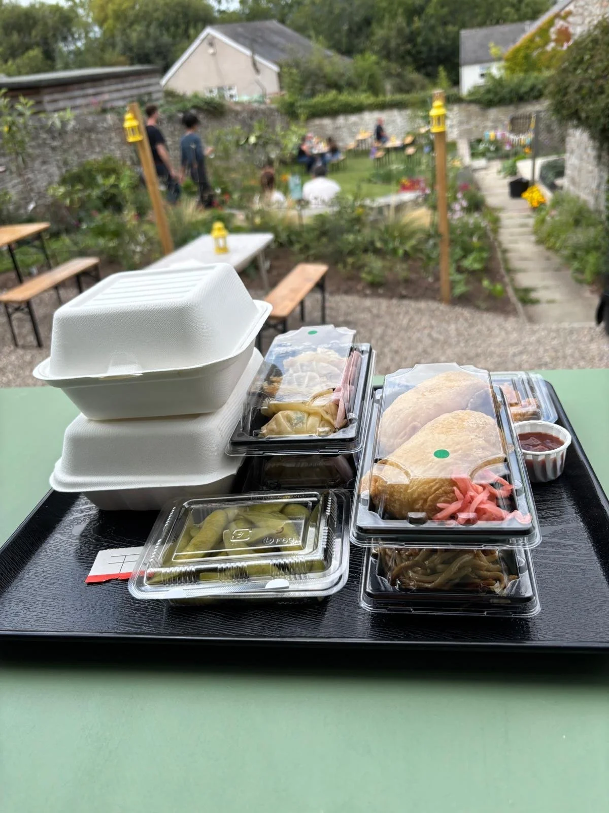 A tray of takeout food including two wrapped burritos, a container of pickled vegetables, a container of green beans, and two foam containers, set on a table with an outdoor garden in the background.
