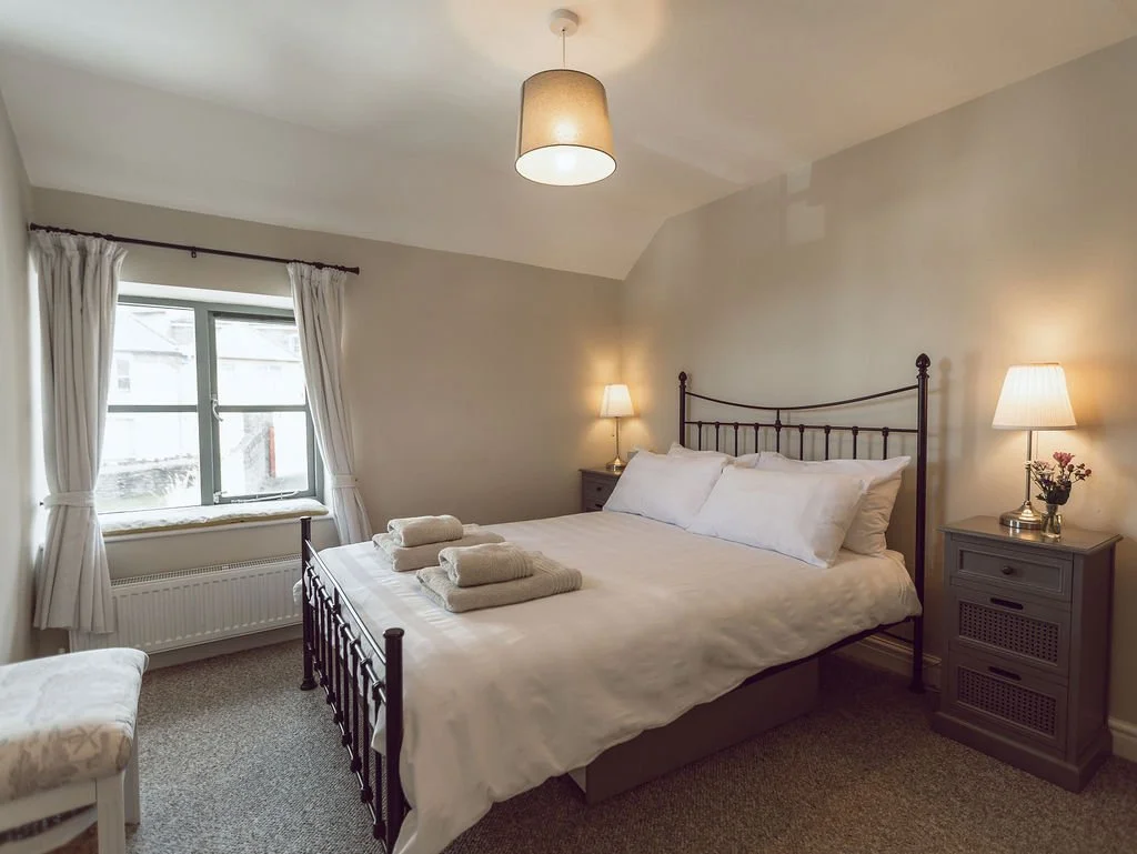 A cozy bedroom with a large bed, white bedding, and folded towels on top. There are two bedside tables with lamps, a window with white curtains, and a small cushioned bench near the window. The room has beige walls and a ceiling light.