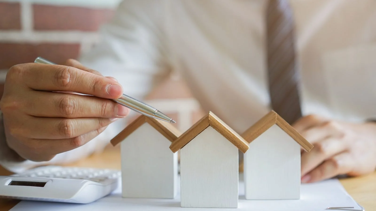 Person holding pen near small wooden house models on desk with calculator.
