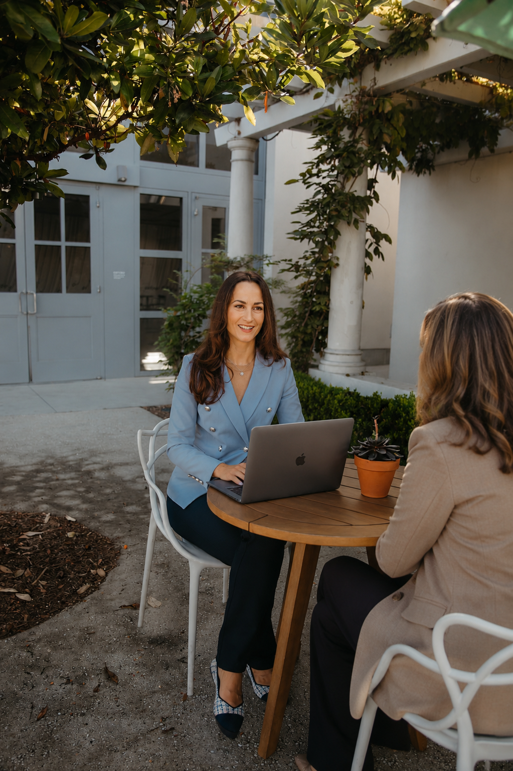 Two women sitting at an outdoor table during daytime, one smiling at the other, with a laptop and a potted plant between them, surrounded by greenery and trees.