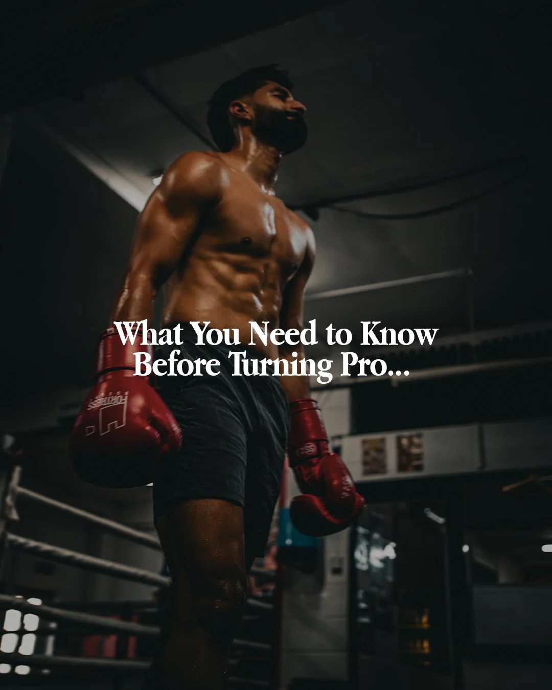 A muscular man with a beard, shirtless and sweating, wearing black shorts and red boxing gloves, standing in a dimly lit boxing gym.