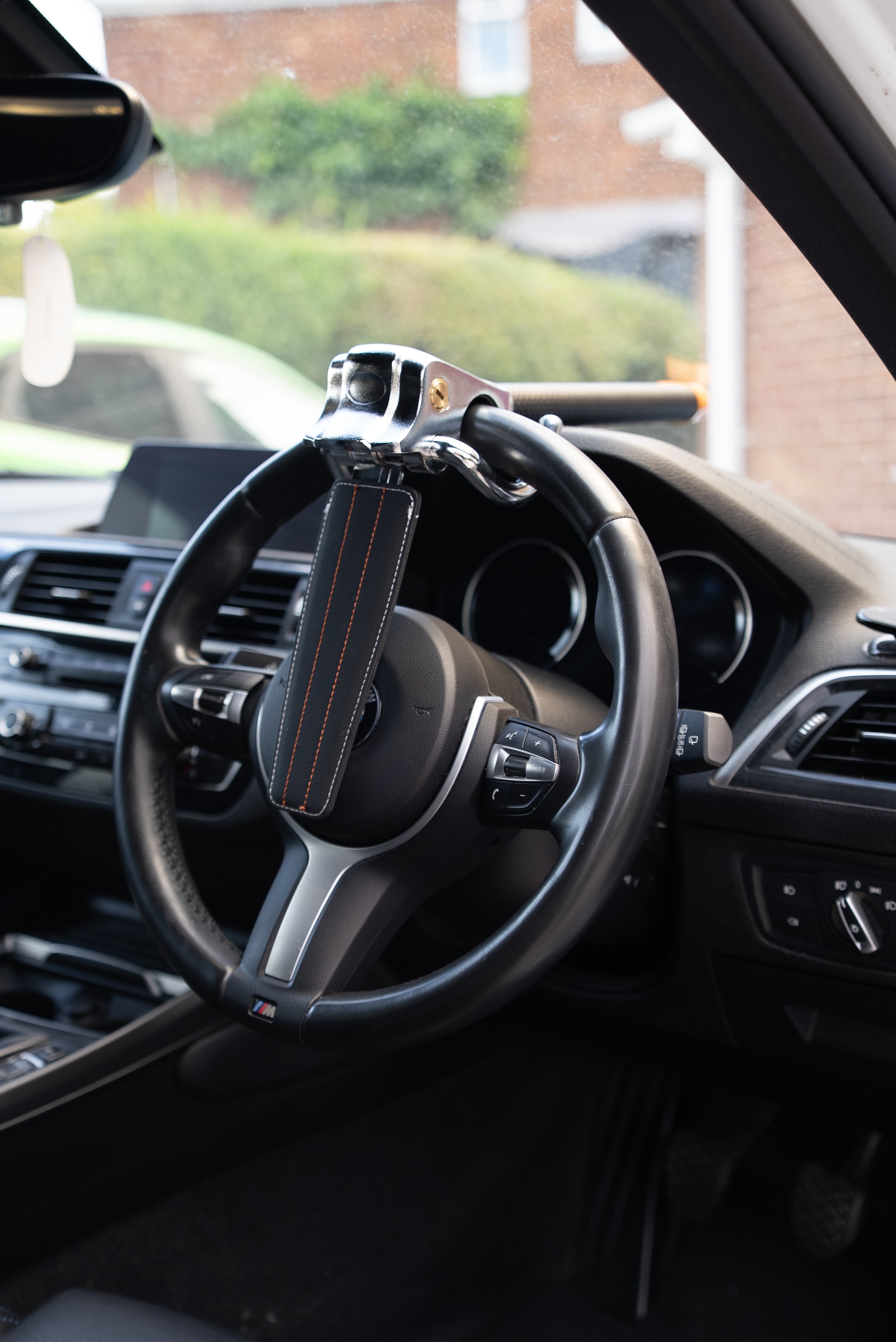 The interior of a car showing a manual steering wheel with BMW M badge, an aftermarket steering wheel lock, and dashboard controls.