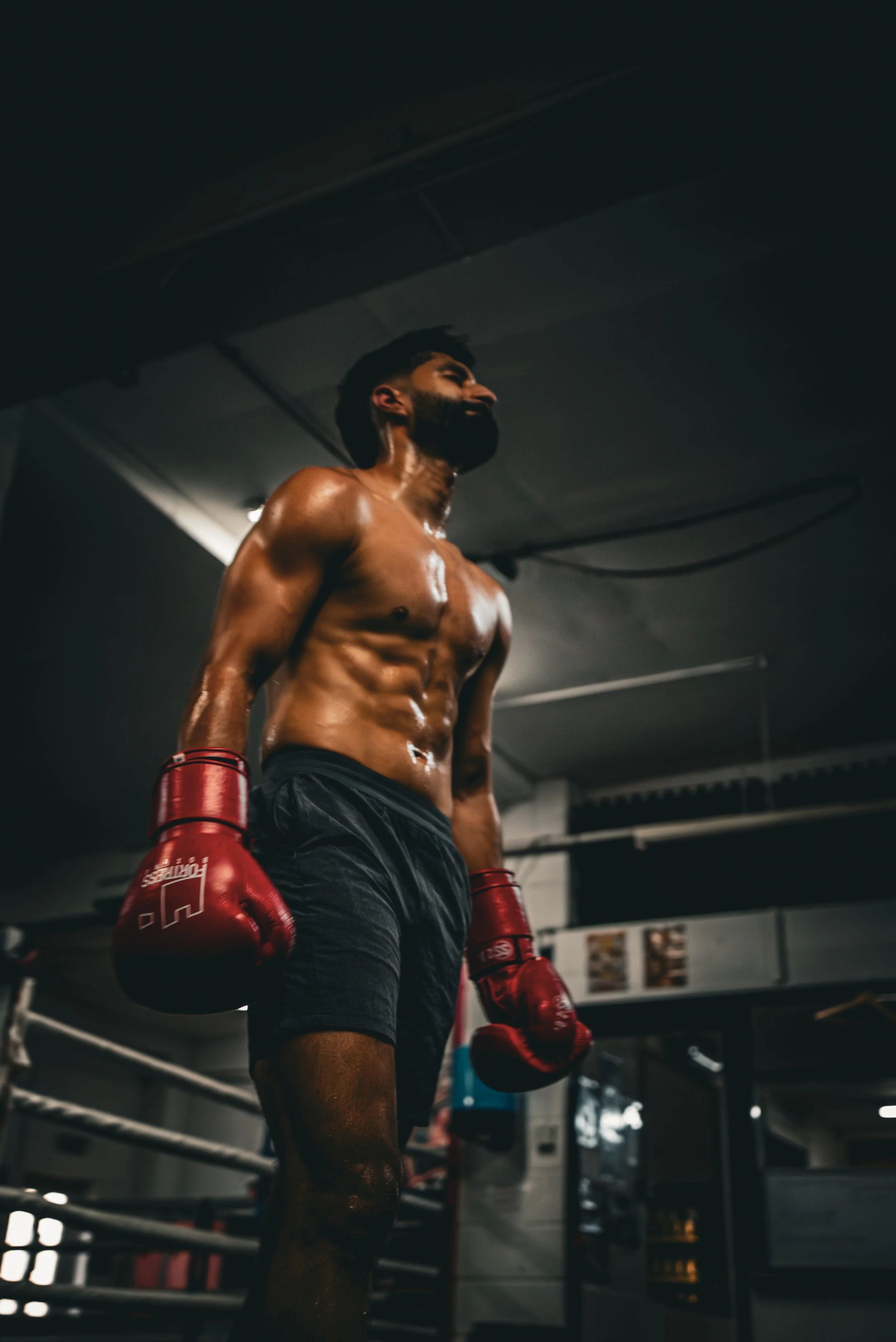 A male boxer with a muscular, sweaty upper body standing in a boxing gym, wearing red boxing gloves and black shorts.