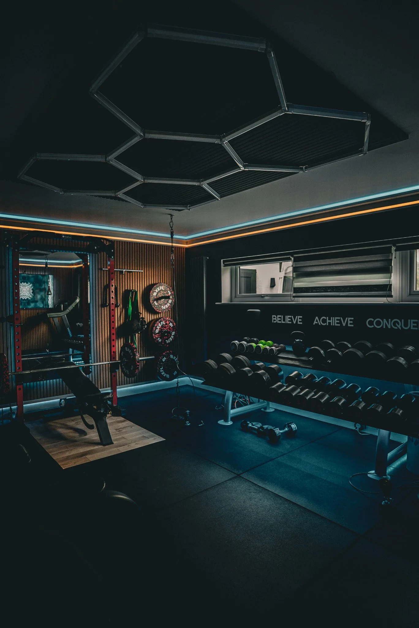 A modern home gym with dumbbells, a bench, weight plates, and a pull-up station, with motivational words on the wall.