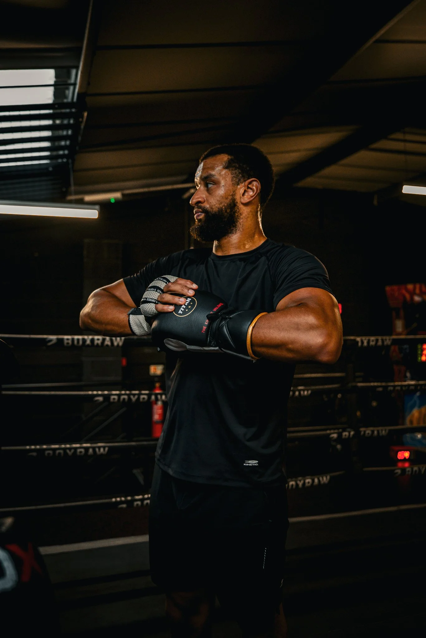 A male boxer in black workout clothes and boxing gloves standing in a boxing gym, looking to the side with his arms crossed.