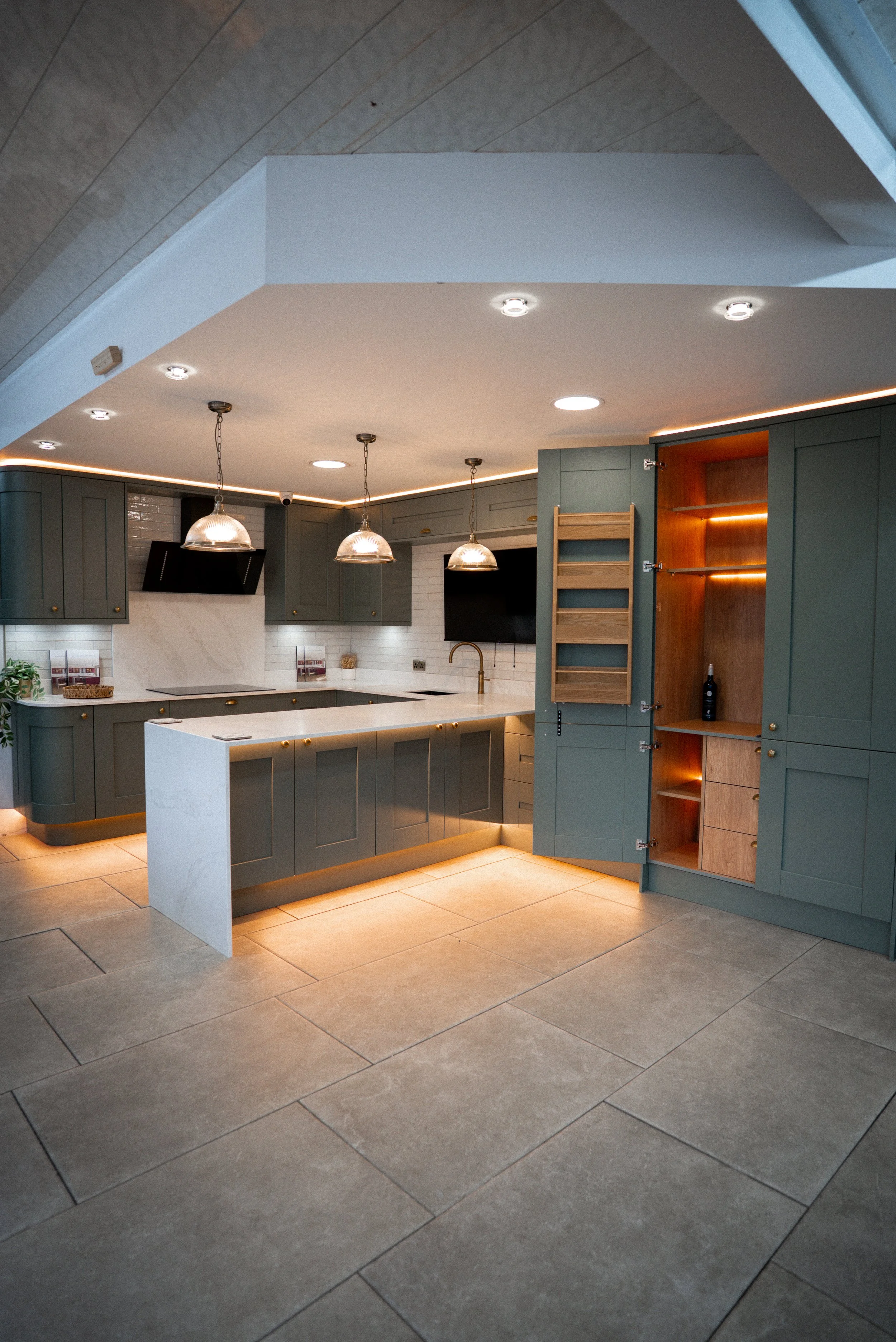 Modern kitchen with gray cabinets, white countertops, pendant lighting, a black range hood, and built-in shelving with interior lighting.