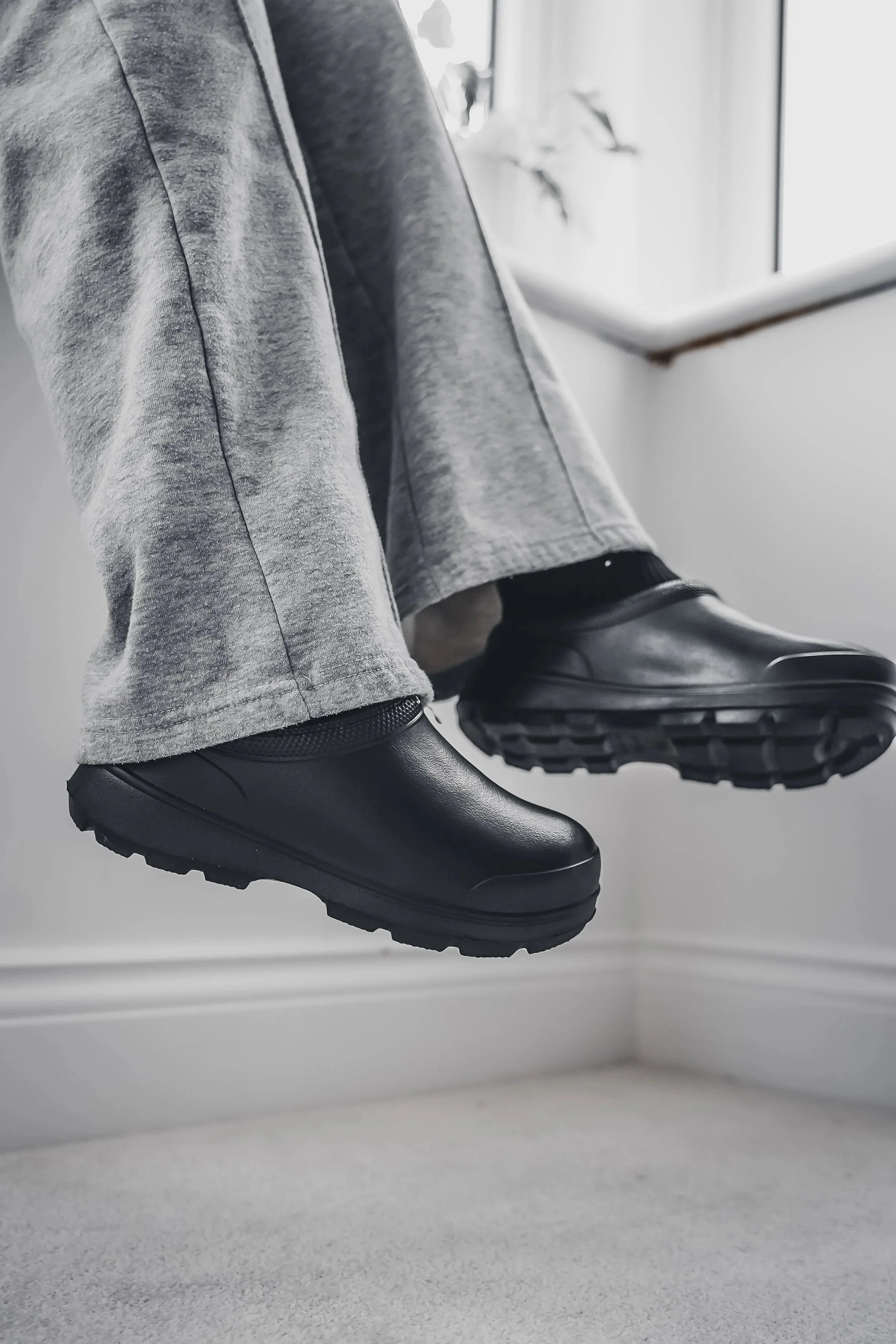 Close-up of a person's feet wearing black waterproof shoes, hanging in front of a window with light grey pants