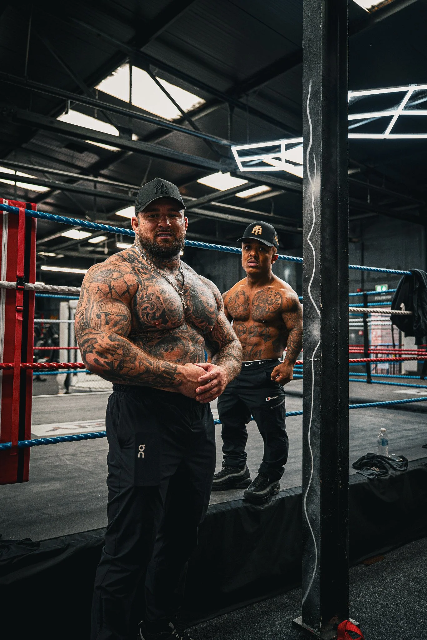 Two shirtless men with tattoos standing in a boxing gym near a boxing ring.