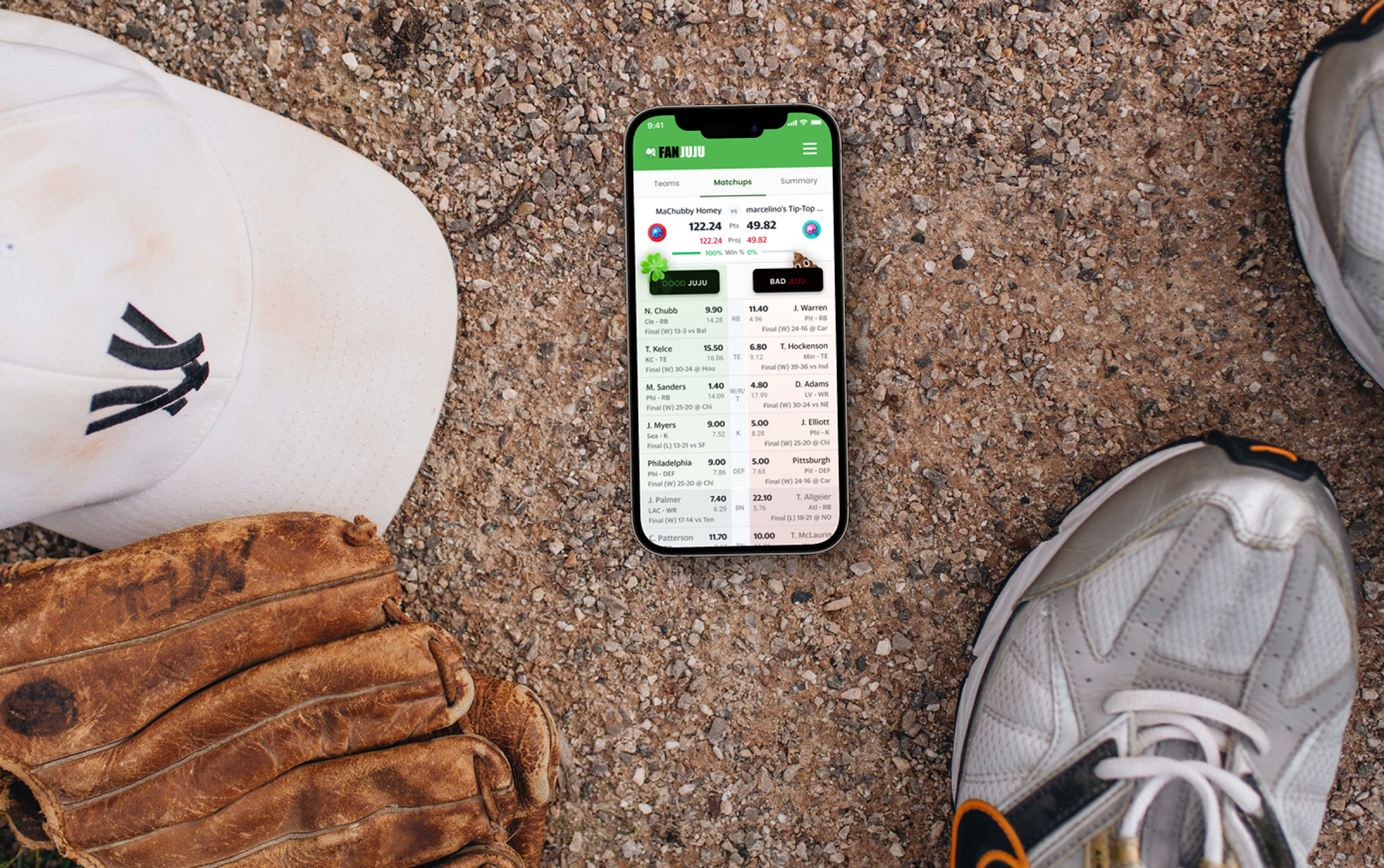 Sports-related flat lay with a smartphone displaying a fantasy sports app, a white baseball cap, a worn baseball glove, and a pair of athletic shoes on a gravel surface.