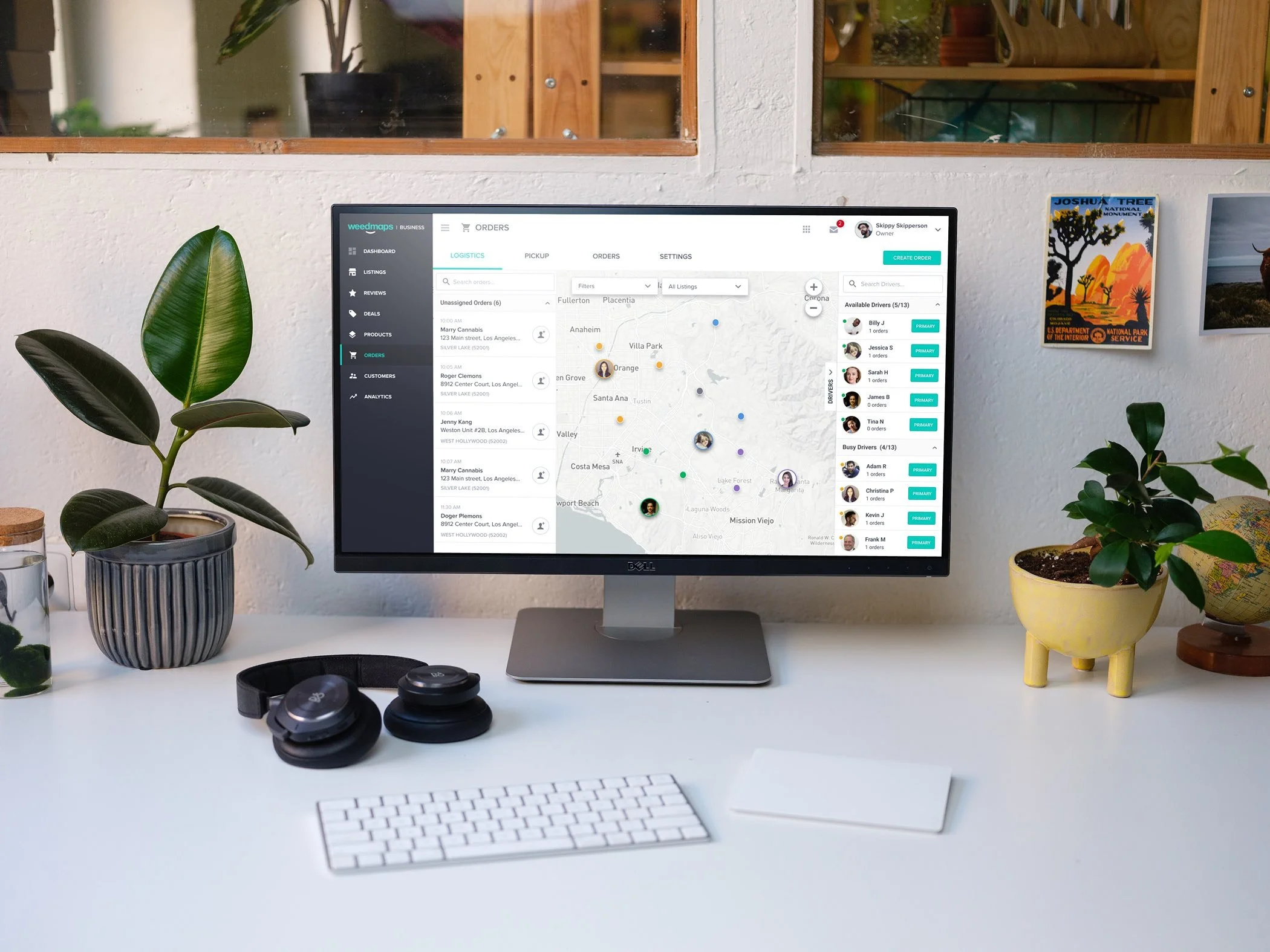 Workspace with a computer monitor displaying logistics software, keyboard, headphones, potted plants, and wall decor.