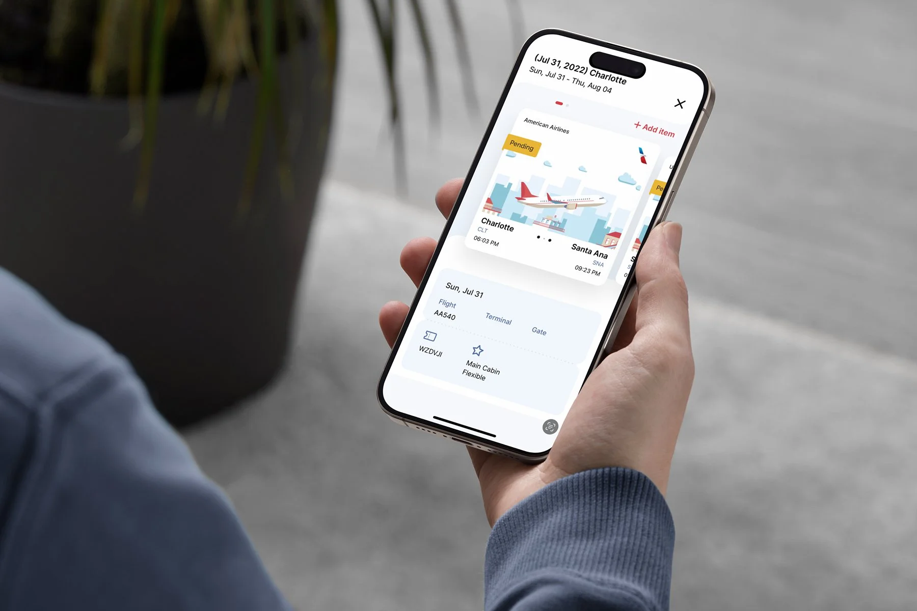 Person holding a smartphone displaying a flight itinerary, showing a flight from Charlotte to Santa Ana on July 31, 2022, with details like airline, flight number, and cabin class.