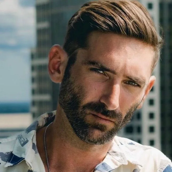 Close-up of a man with a beard and styled hair standing outdoors, wearing a patterned shirt.
