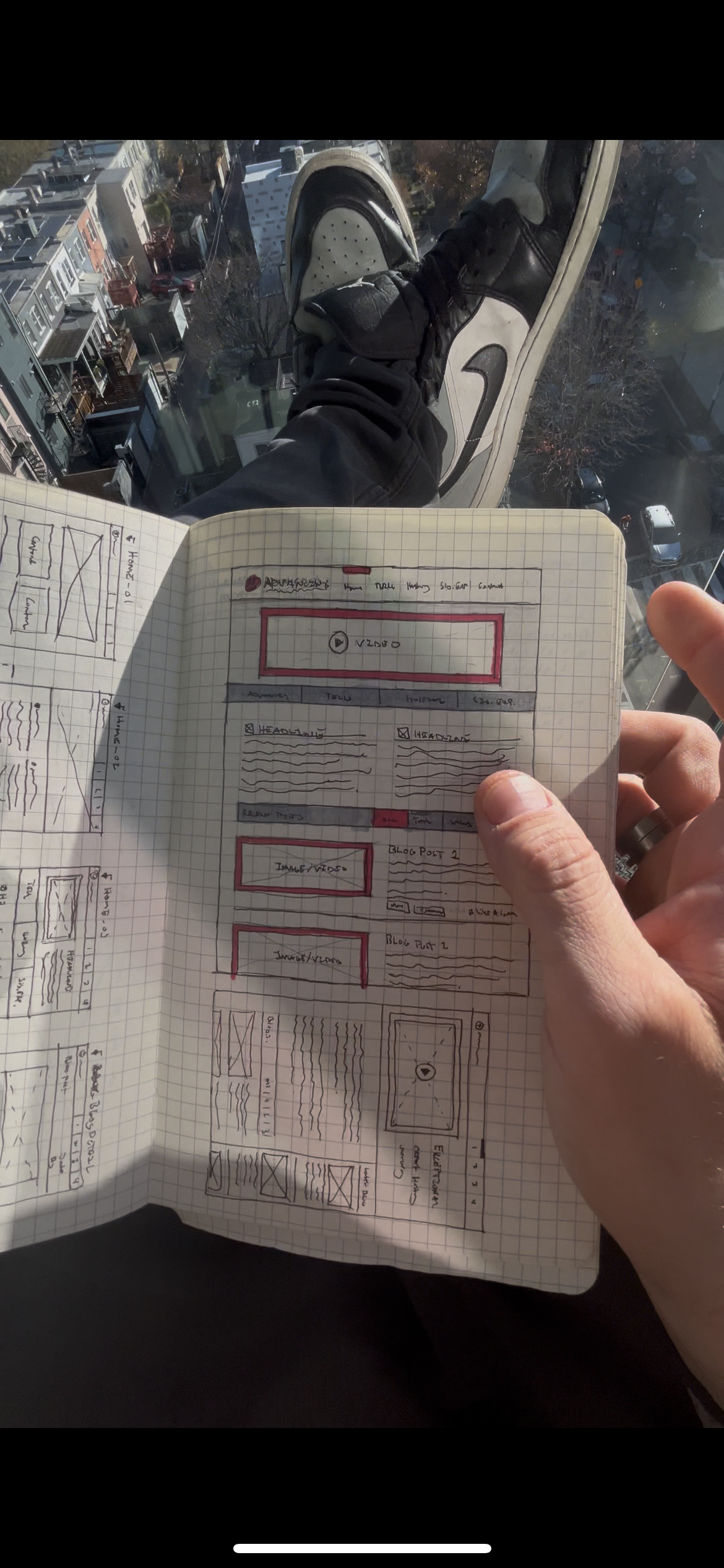 Person holding a notebook with sketches of website wireframes, dressed in sneakers, with a city view in the background.