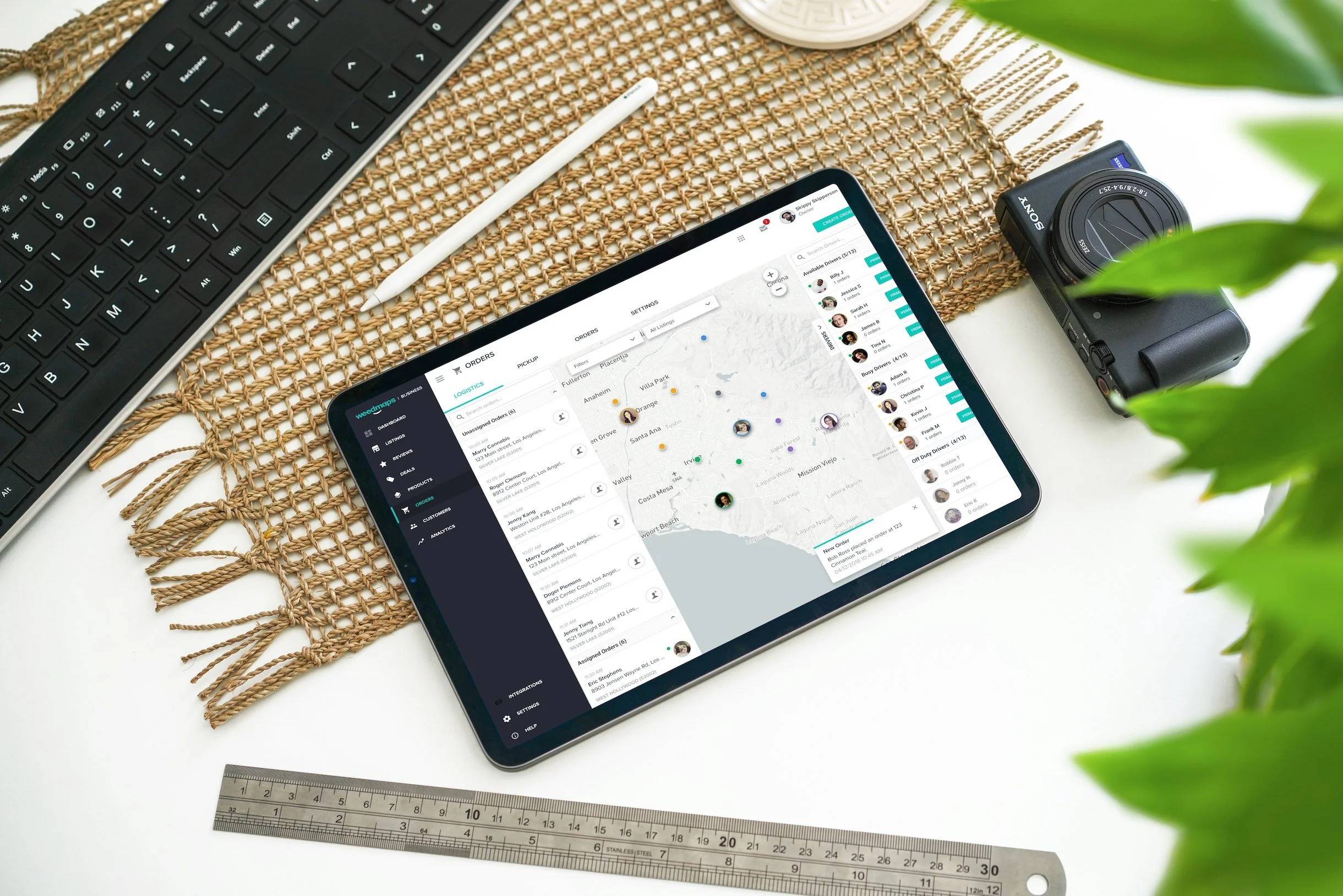 Tablet displaying map and lists on a woven mat, next to keyboard, camera, pen, ruler, and plant.