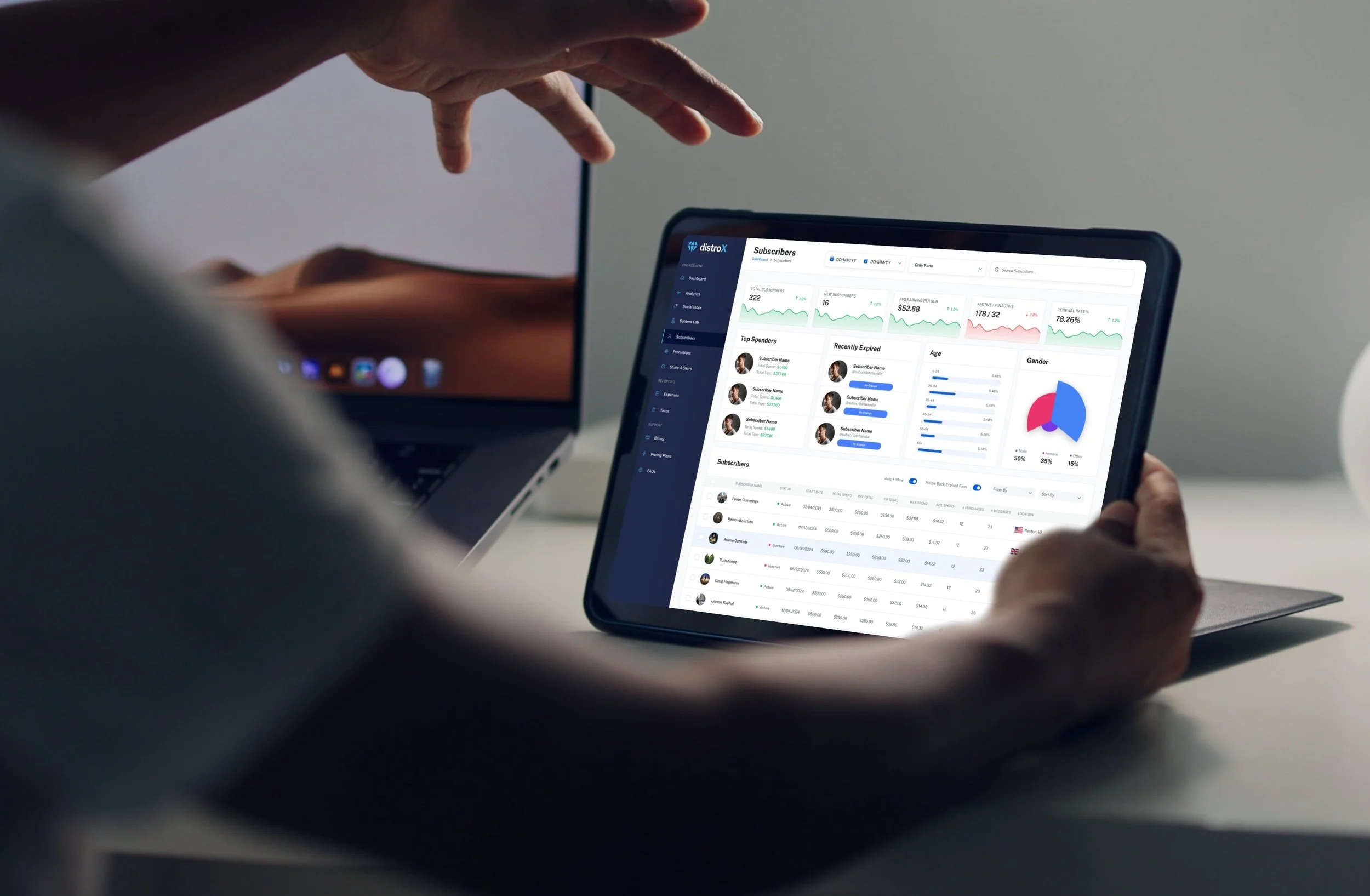 Person viewing a tablet displaying a data dashboard with graphs, charts, and subscriber information on a desk next to a laptop.
